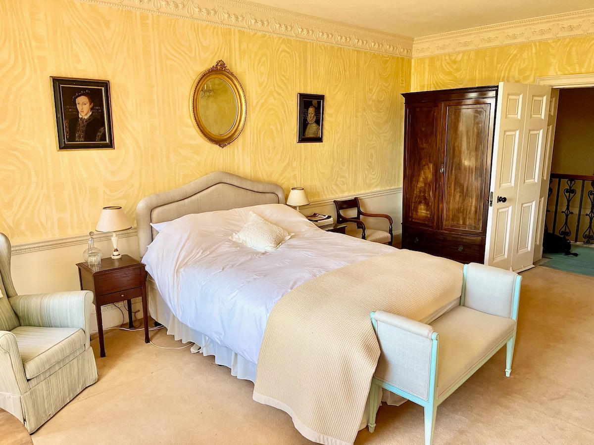 A room features soft yellow walls and a spacious bed dressed in white linens with a decorative cushion. Two armchairs are positioned next to a small bedside table. A vintage wardrobe stands against one wall, while a door leads to another area.