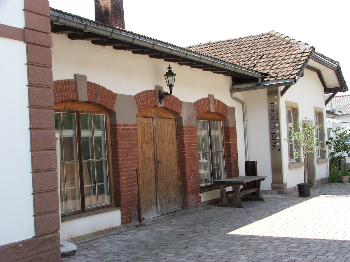 The exterior of a single-story building features a warm color palette of white and brick accents. Large windows with multiple panes provide natural light. A sturdy wooden door, flanked by two lanterns, leads into the interior. A rustic wooden table is positioned nearby on the cobblestone surface.