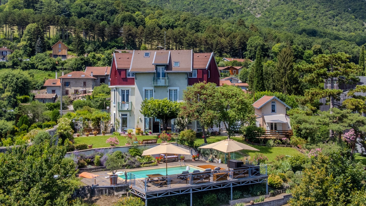 A spacious independent house is surrounded by greenery and mountains. The property features a private pool with lounge chairs and umbrellas, set in a beautifully landscaped garden. Various rooftops of nearby residences and lush trees create a serene backdrop against the mountainous landscape.