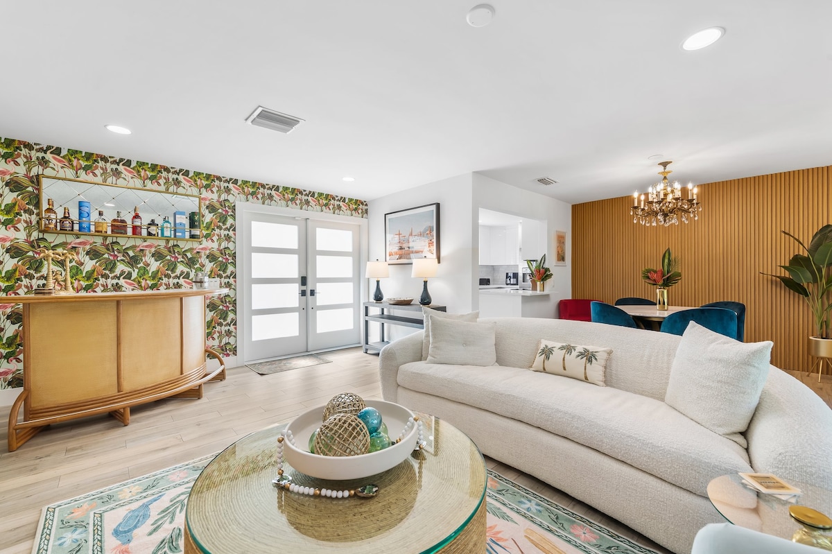 A stylish living area features a light-colored sofa with decorative pillows, complemented by a round coffee table showcasing decorative glass objects. A vibrant foliage wallpaper adorns one wall, while a chic bar cart is visible in the background, adding a touch of elegance to the space.