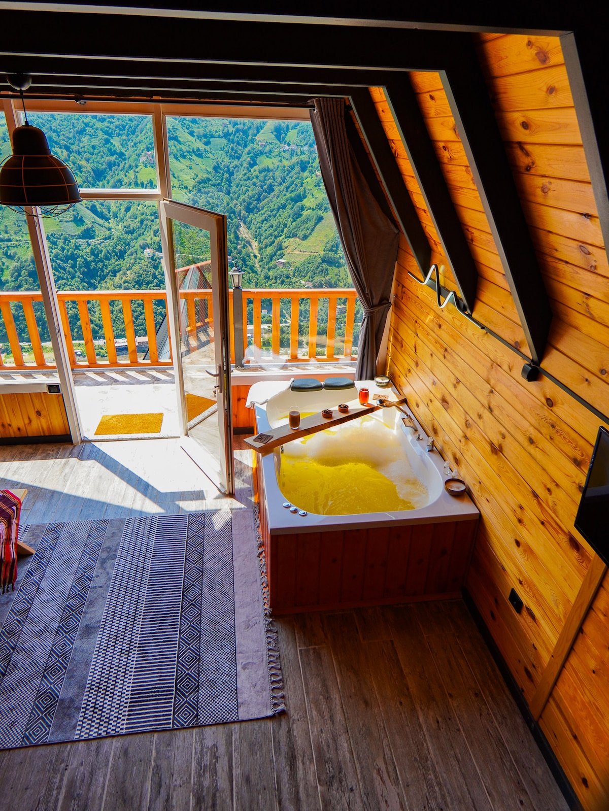 A spacious interior features a wooden decor with an open balcony showcasing lush green landscapes. A hot tub sits invitingly beside the window, filled with warm water. The area is softly illuminated, complemented by a patterned rug on the wooden floor.