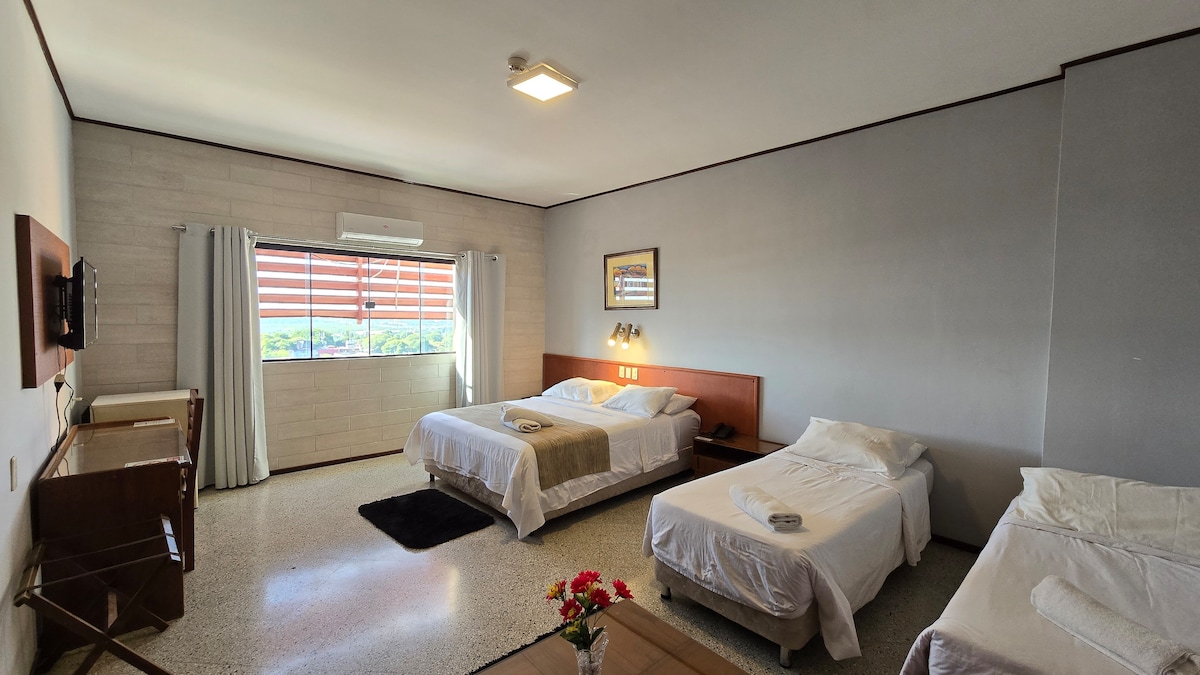 A spacious guest room is presented, featuring two large beds and one single bed arranged along the walls. A wooden dresser and a flat-screen television are visible. Natural light filters through the window, complemented by light-colored curtains. A small flower arrangement on a table adds a touch of color.