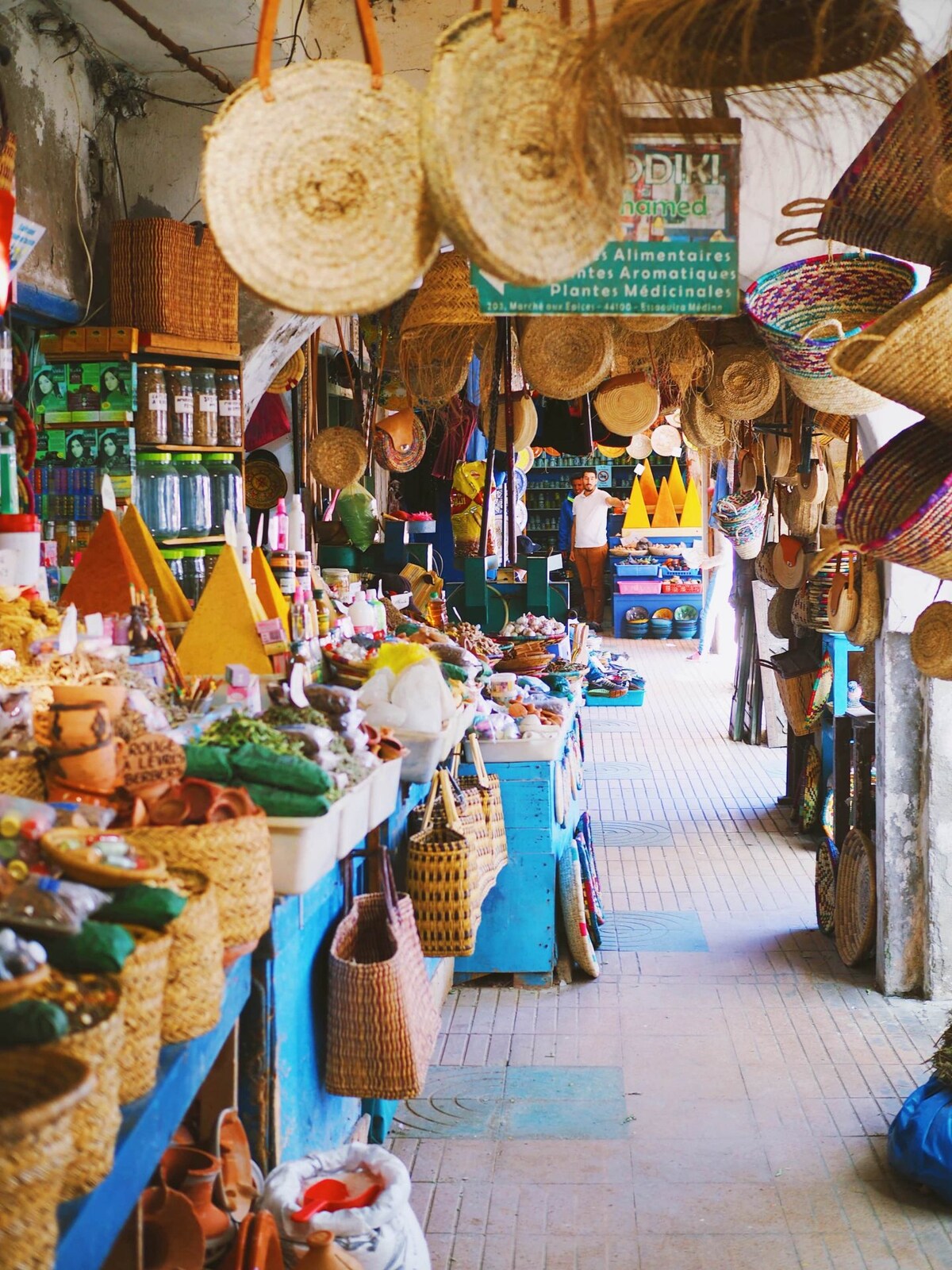 A vibrant market scene is captured, filled with various woven baskets and colorful hats. Shelves display an array of items, contributing to a lively atmosphere. The space is illuminated by natural light, enhancing the texture and colors of the local crafts on display.