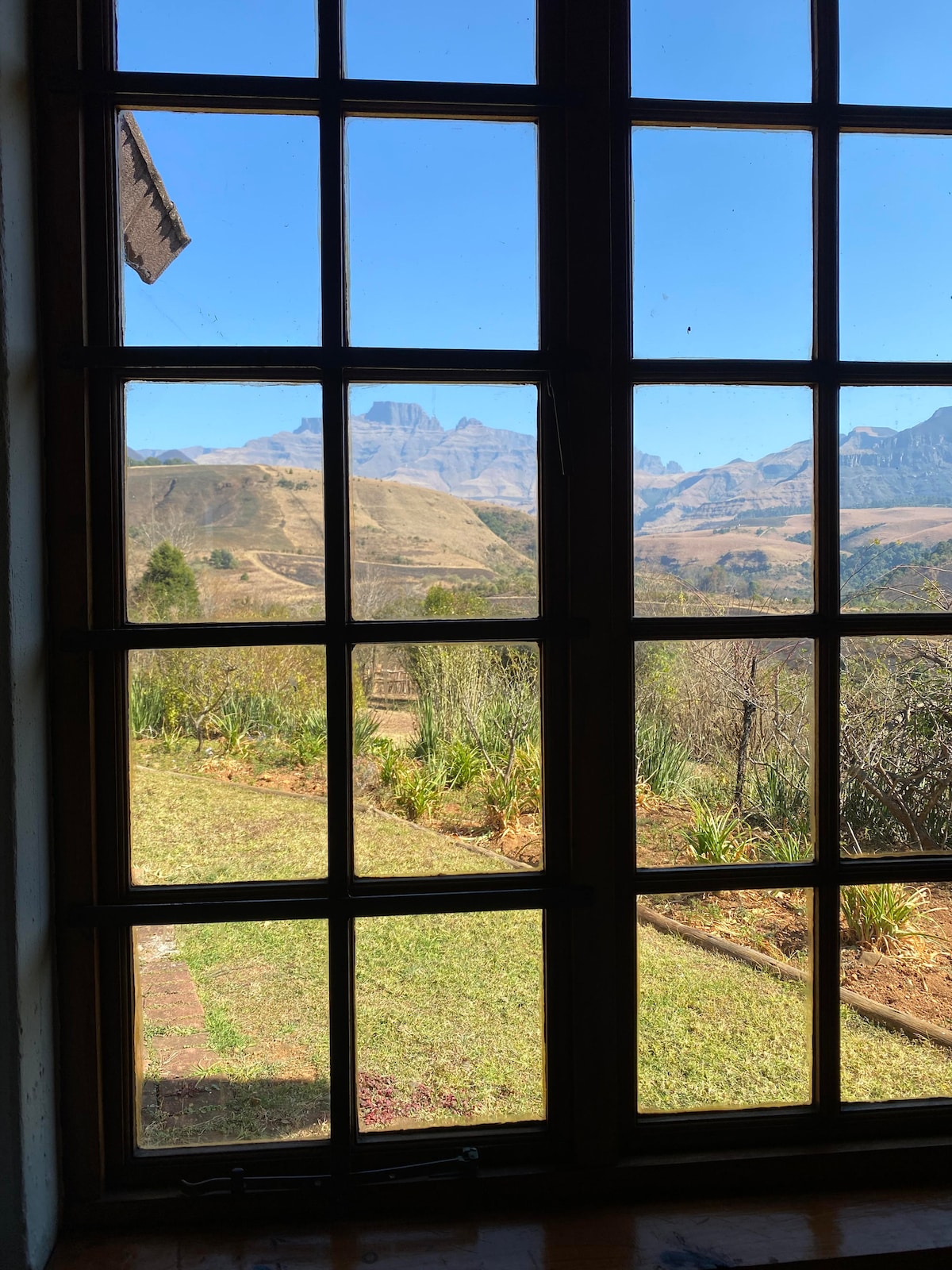 A large window offers a clear view of the Drakensberg mountains, framed by multiple panes. The lush greenery in the foreground complements the natural landscape, creating a sense of openness and connection to the outdoors.