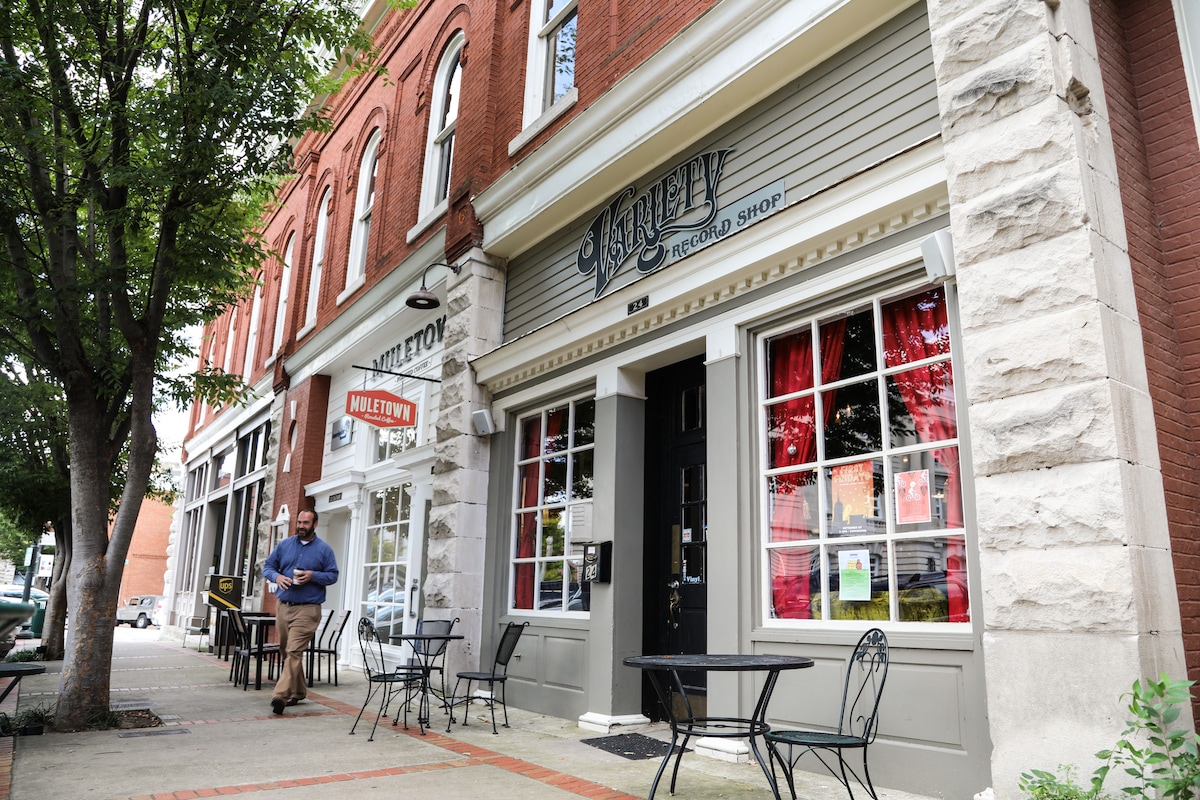 The image depicts a charming storefront with distinctive architecture, featuring large windows draped in red curtains. Outdoor seating, consisting of small, round tables and black chairs, lines the sidewalk. A pedestrian is shown walking by, capturing the vibrant atmosphere of the downtown area.