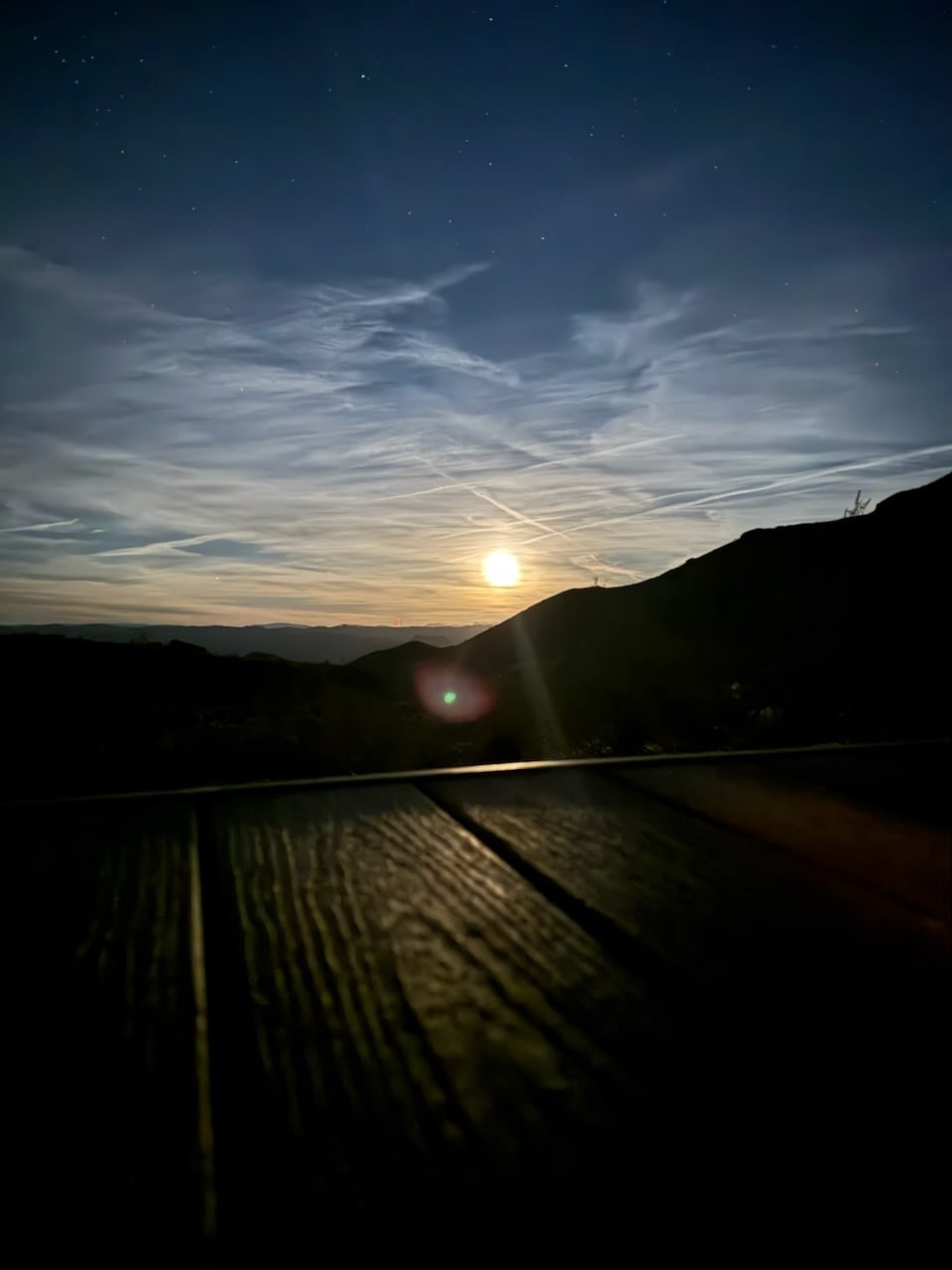 A serene twilight landscape unfolds where soft clouds are illuminated by a glowing moon above mountain silhouettes. The wooden deck is visible in the foreground, inviting moments of relaxation while enjoying the peaceful view.