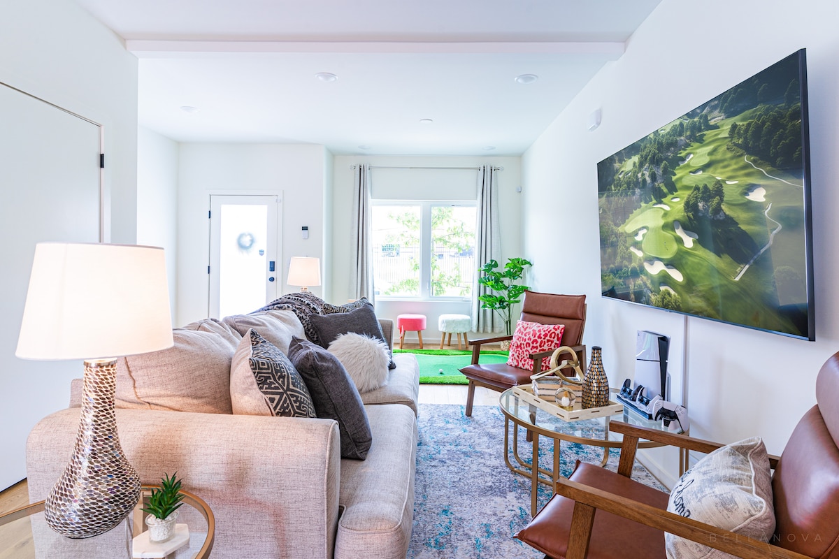 A bright living room is furnished with comfortable seating, including a beige sofa and brown armchairs. A large television displaying a scenic golf course is mounted on the wall. Natural light flows in through the window, enhancing the inviting ambiance of the space.