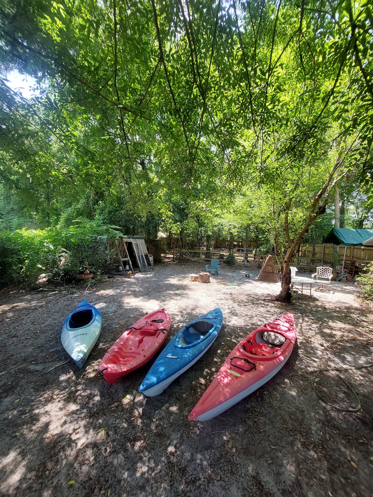 Four kayaks are arranged on a sandy surface beneath a canopy of trees, creating a shaded area. A play area for animals is visible in the background, with seating options nearby. The serene environment emphasizes a connection to nature.