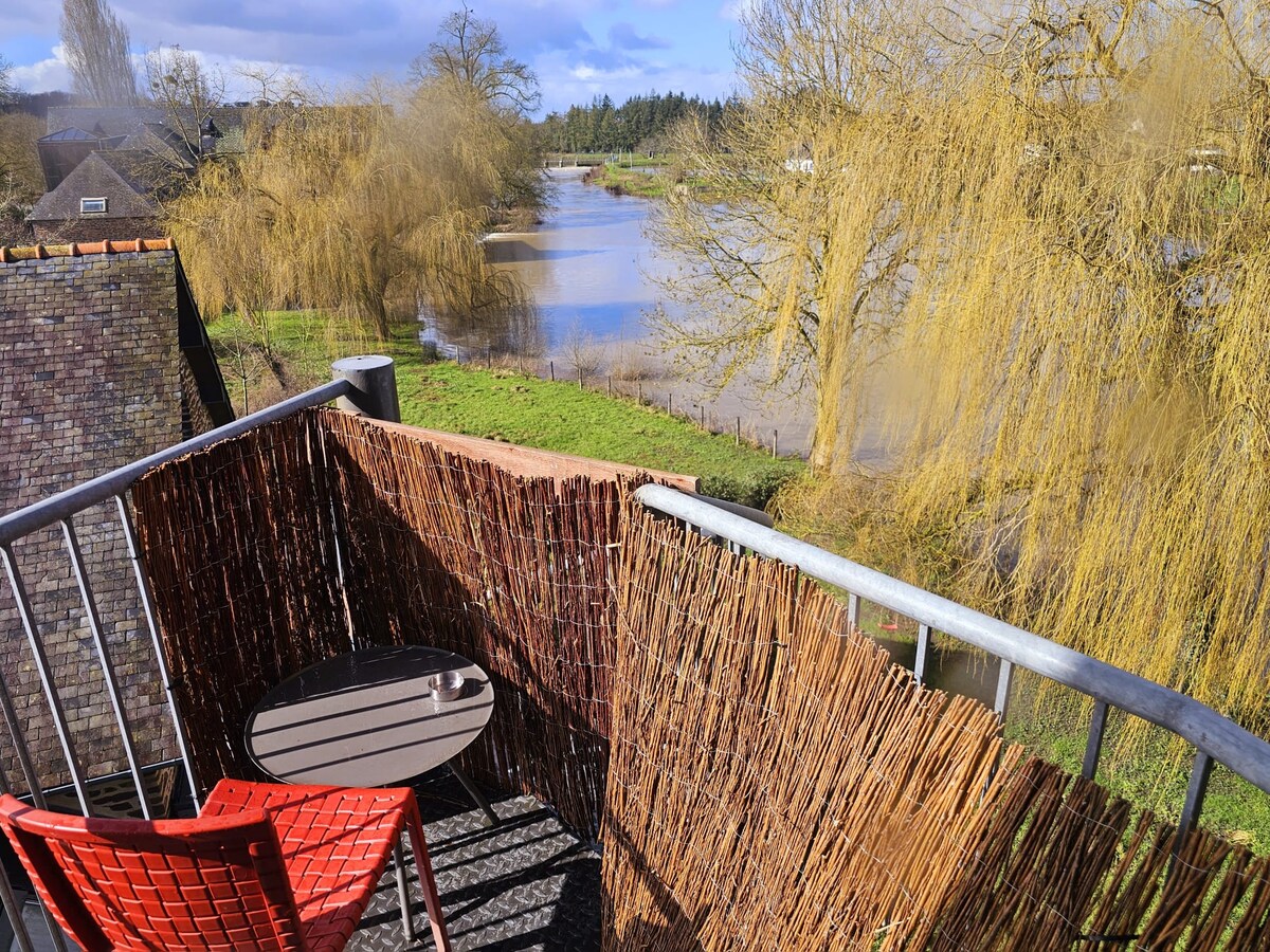 A private balcony features a round table and a single red chair, overlooking a serene river framed by willow trees. The setting offers a peaceful view of the lush greenery and water, providing a quiet space for relaxation.