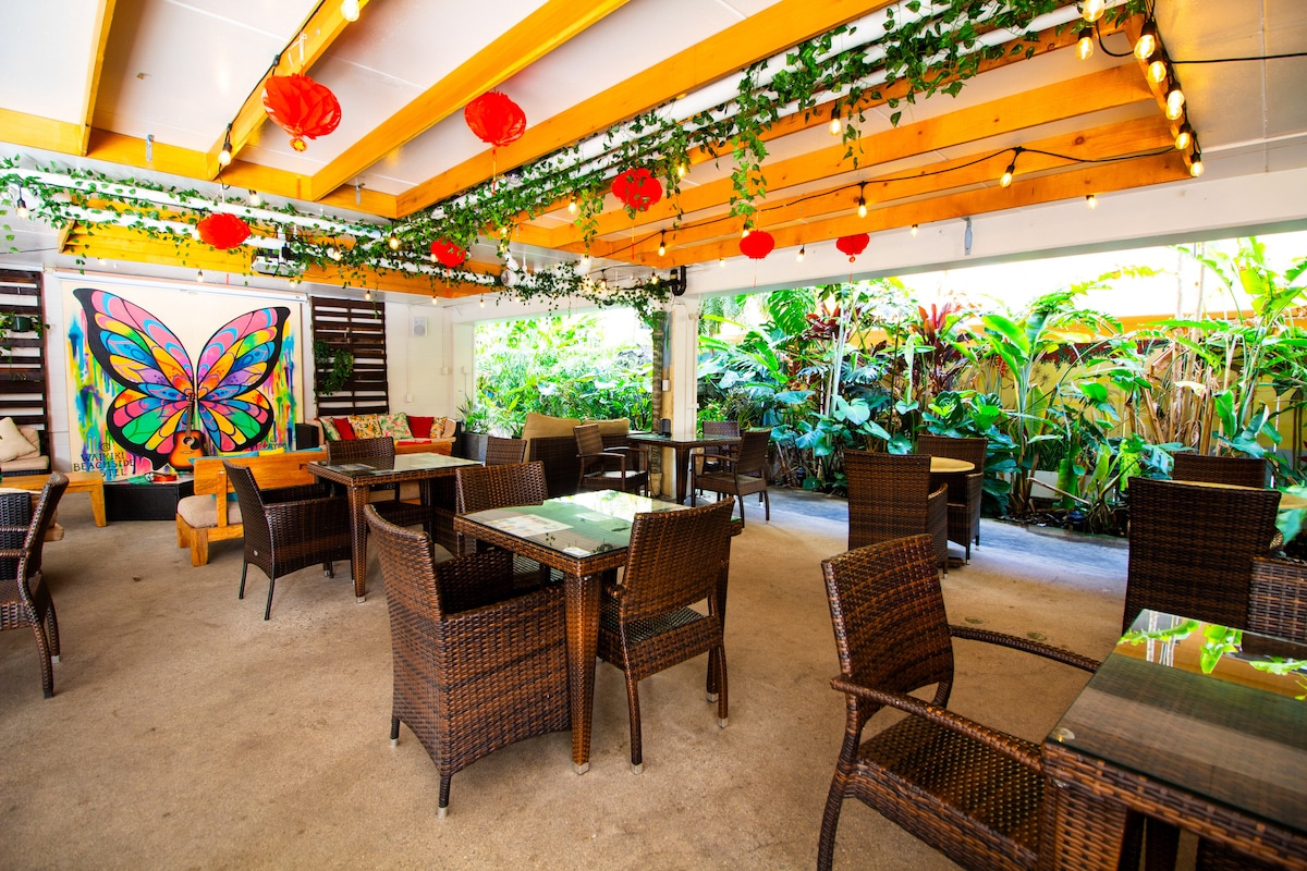 An outdoor seating area is filled with wicker chairs and tables, surrounded by lush greenery. A vibrant butterfly mural is displayed on the wall, complemented by string lights and decorative paper lanterns hanging from the wooden beams above.