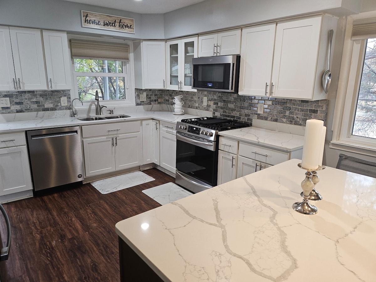A sleek kitchen is presented with white cabinetry and a contrasting marble countertop. Stainless steel appliances, including a gas stove and dishwasher, are visible. A window allows natural light to fill the space, highlighting the modern backsplash and decorative accents.