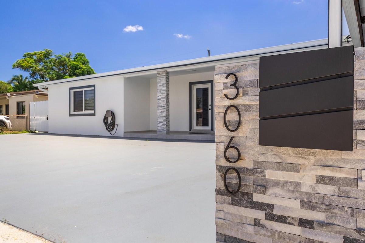 The exterior of the apartment is showcased, featuring a modern design with a stone accent wall. A bold house number is displayed prominently on the stone facade. A clean driveway leads to the entrance, framed by lush greenery under a clear blue sky.
