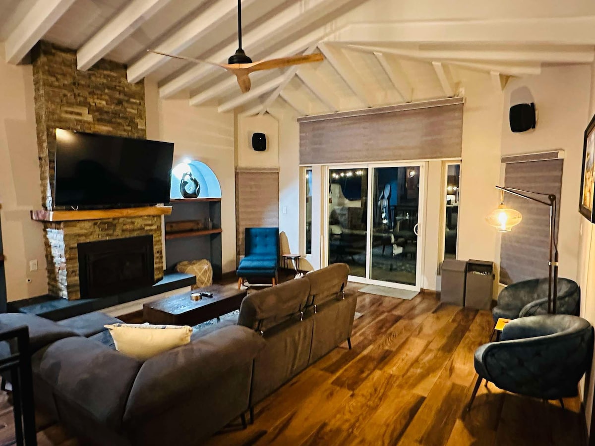 A spacious living room features high ceilings with exposed beams and wood flooring. A large television is mounted above a stone fireplace. Comfortable seating is arranged around a central coffee table, with natural light streaming through the sliding glass doors leading to the patio.
