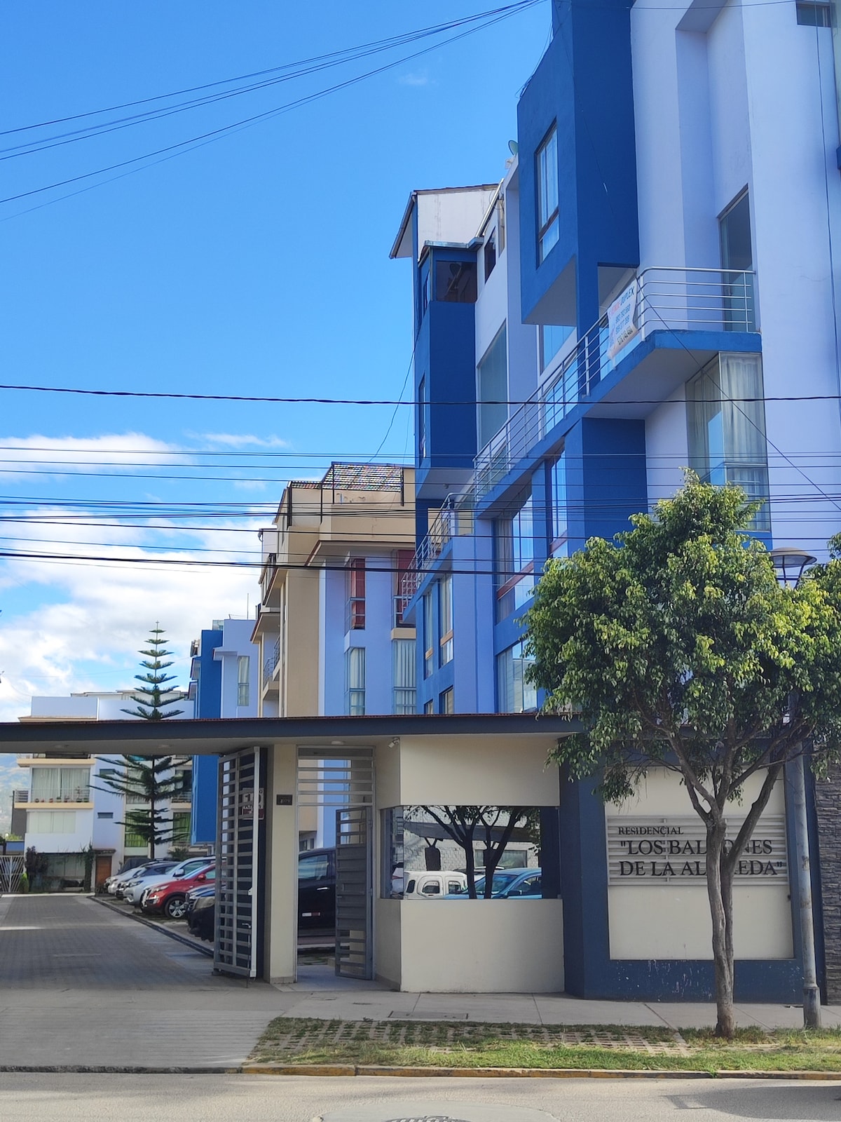 A modern building is visible, featuring a combination of blue and white hues. The entrance is marked by a secure gate, with multiple floors of windows allowing natural light inside. Vehicles are parked nearby, with greenery surrounding the entrance, enhancing the inviting exterior.