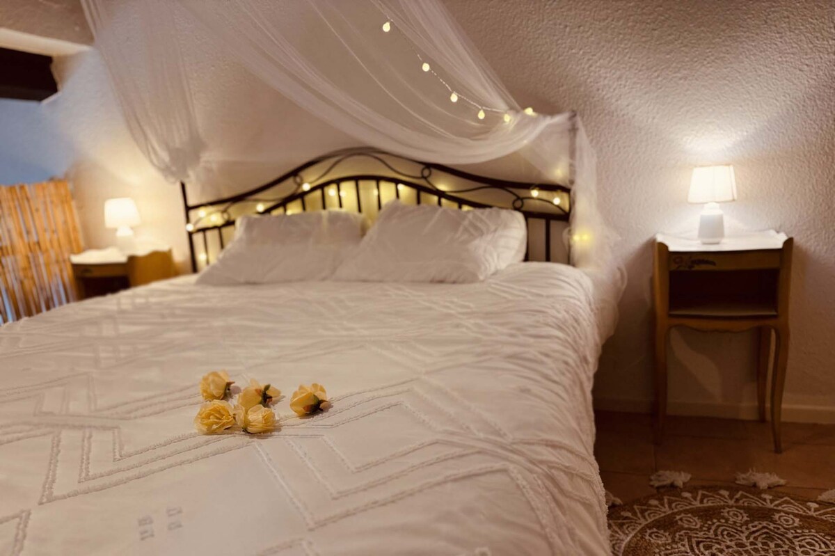 A inviting sleeping area features a spacious bed dressed in white linens, accentuated by soft fairy lights overhead. A delicate arrangement of yellow flowers lies on the bed, with a small bedside table and lamp providing additional ambient lighting for comfort.