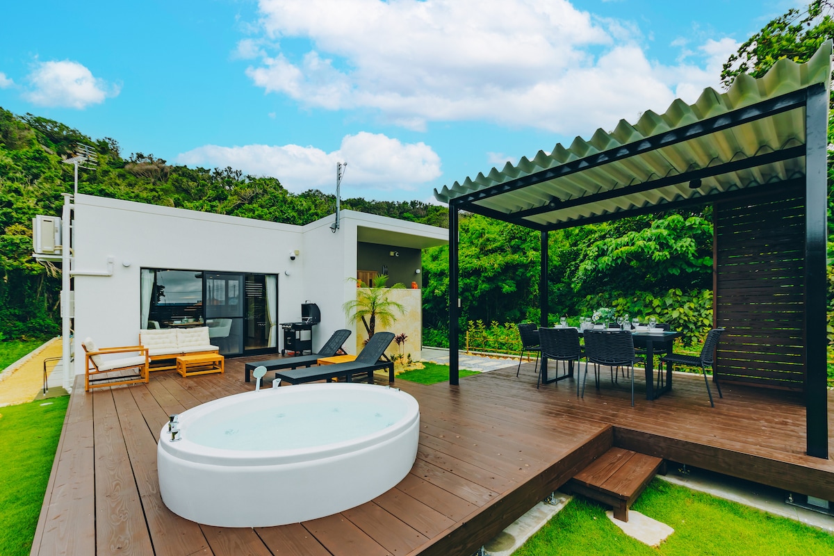 An outdoor area features a modern, private jacuzzi surrounded by a spacious wooden deck. A shaded dining area is equipped with a large table and chairs, while sun loungers are positioned nearby. Lush greenery provides a serene backdrop.