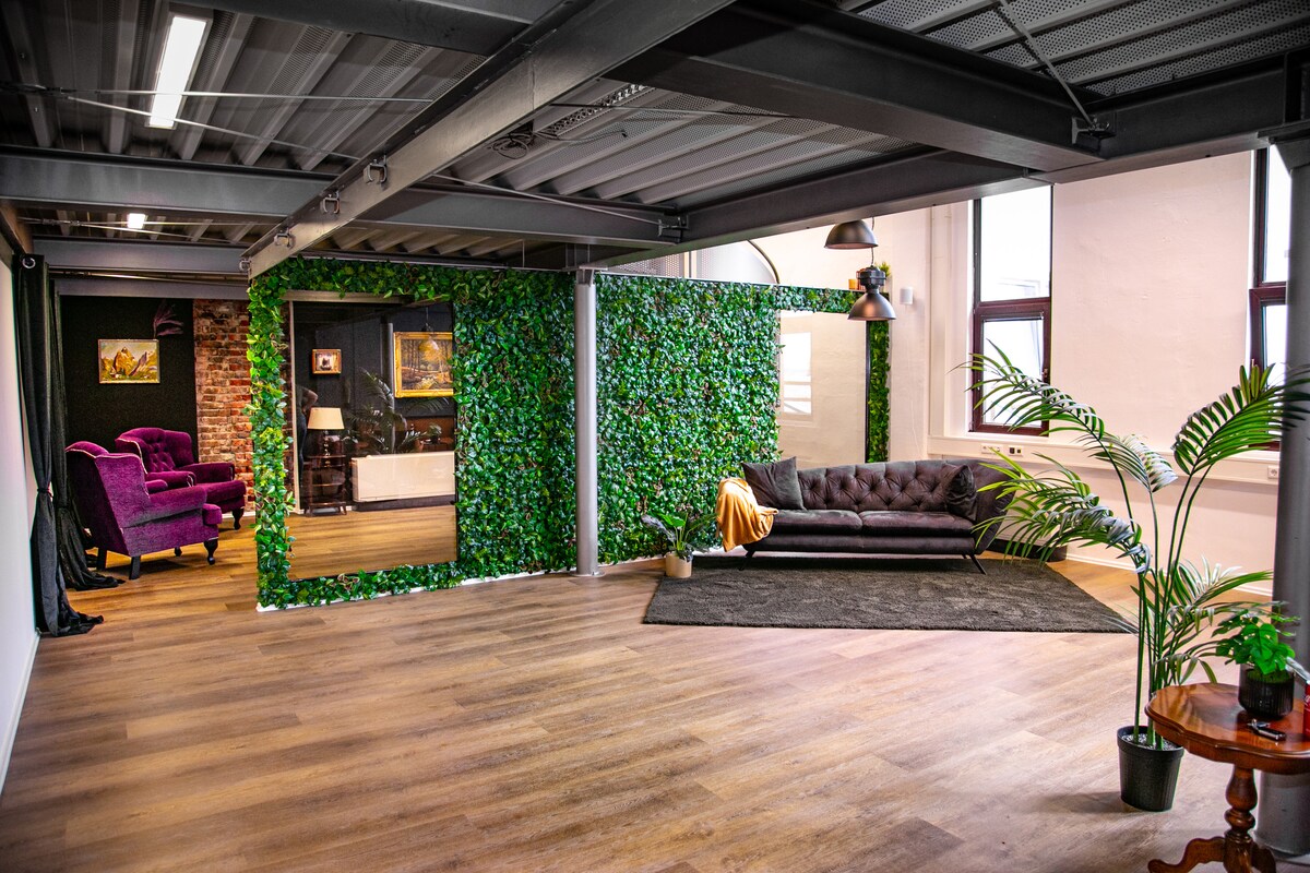 A spacious room features a green wall adorned with artificial foliage, contrasting with the industrial-style exposed ceiling. A dark sofa rests on a light wooden floor, accompanied by a cozy area rug. Natural light enters through large windows, enhancing the inviting layout.
