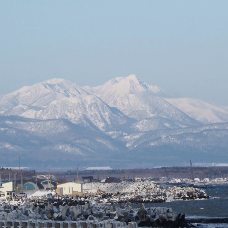 A tranquil coastal scene features snow-capped mountains rising in the distance, contrasting against a clear blue sky. The shoreline is lined with rugged rock formations, creating a natural border along the water's edge. Small structures can be seen dotting the landscape, contributing to the serene atmosphere.