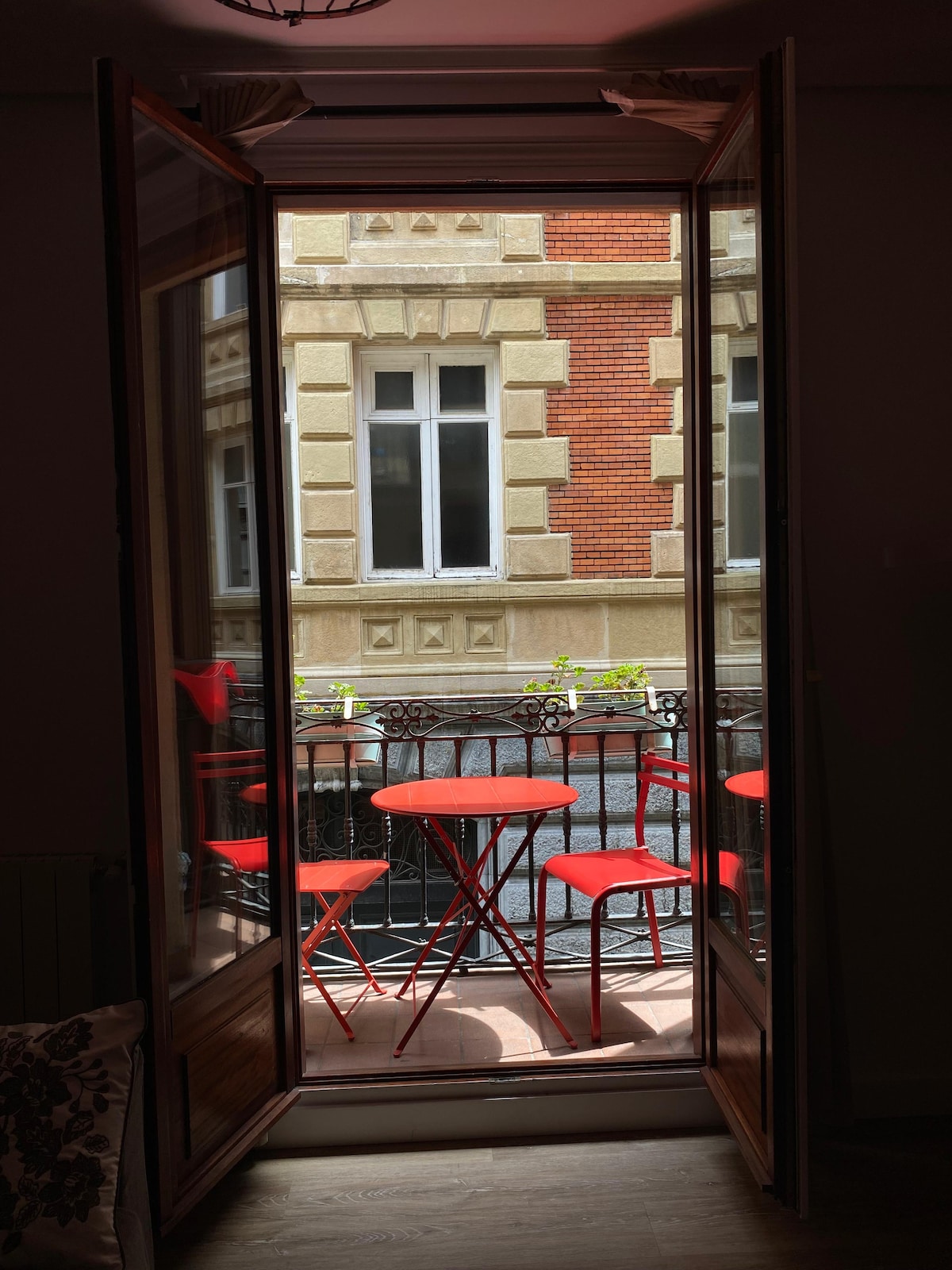 A balcony is shown with two red bistro tables and matching chairs, surrounded by historic brick and stone architecture. Large windows allow natural light to fill the space, while greenery is visible in the distance, providing a refreshing view.