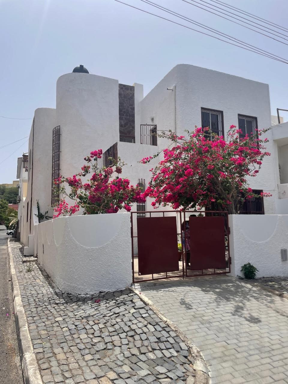 A modern house with a smooth white exterior is surrounded by vibrant bougainvillea, creating a colorful contrast. A gated entrance, painted in red, leads into the property. The pathway is paved with cobblestones, enhancing the charming streetscape.