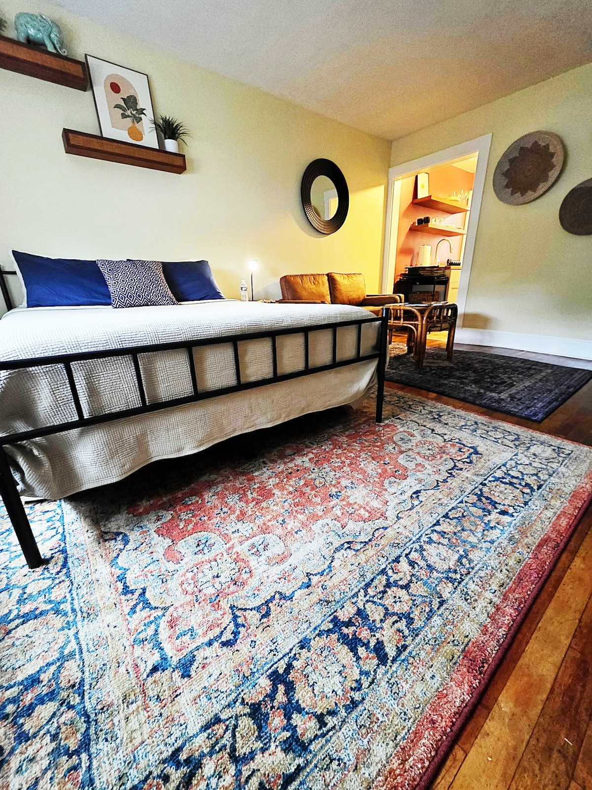 A cozy studio space features a spacious bed with a light-colored blanket, positioned in front of a decorative rug showcasing intricate patterns. A small seating area with a couch is visible, along with a glimpse of the kitchen area through an open doorway.