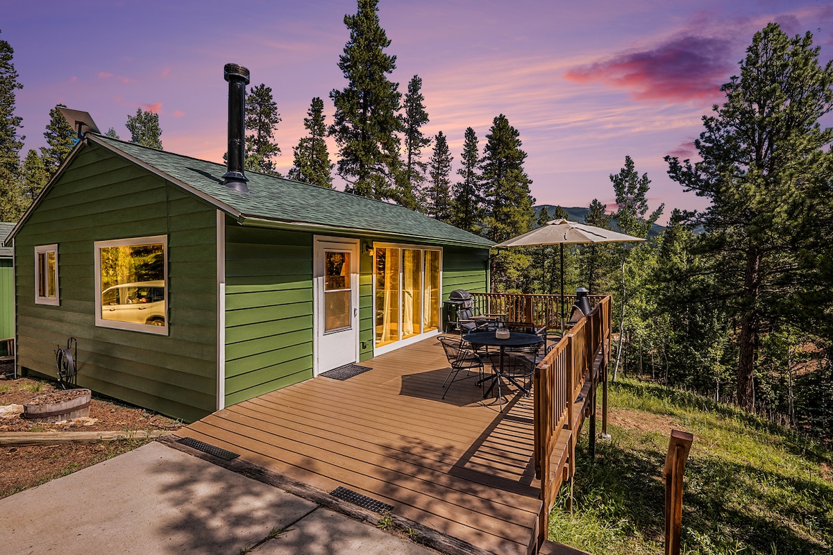 The cabin’s exterior features a green façade and a spacious deck, equipped with a table and chairs. A grill is positioned next to the seating area, surrounded by trees. Large windows allow natural light, creating a connection with the outdoor environment.