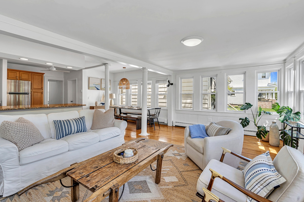 A bright living area is showcased, featuring light-colored furniture and a wooden coffee table. Large windows allow natural light to illuminate the space, which connects to the dining area visible in the background. Potted plants add a touch of greenery to the inviting atmosphere.