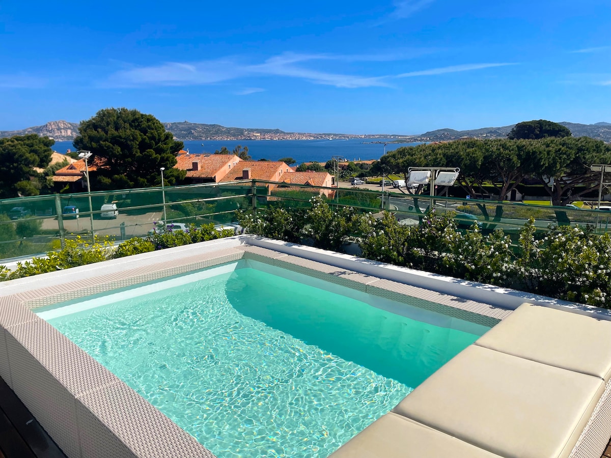 A private heated pool is shown in the foreground, surrounded by a sleek deck. The view features lush greenery and a panoramic backdrop of the sea, with distant coastal hills. Bright blue skies contribute to a serene atmosphere.