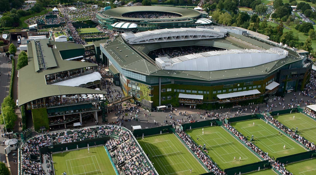 The image presents an aerial view of the Wimbledon Tennis grounds, showcasing multiple tennis courts surrounded by large crowds. The iconic Centre Court is prominently featured, with shaded seating areas visible. Lush greenery surrounds the venue, contributing to the vibrant atmosphere of the event.