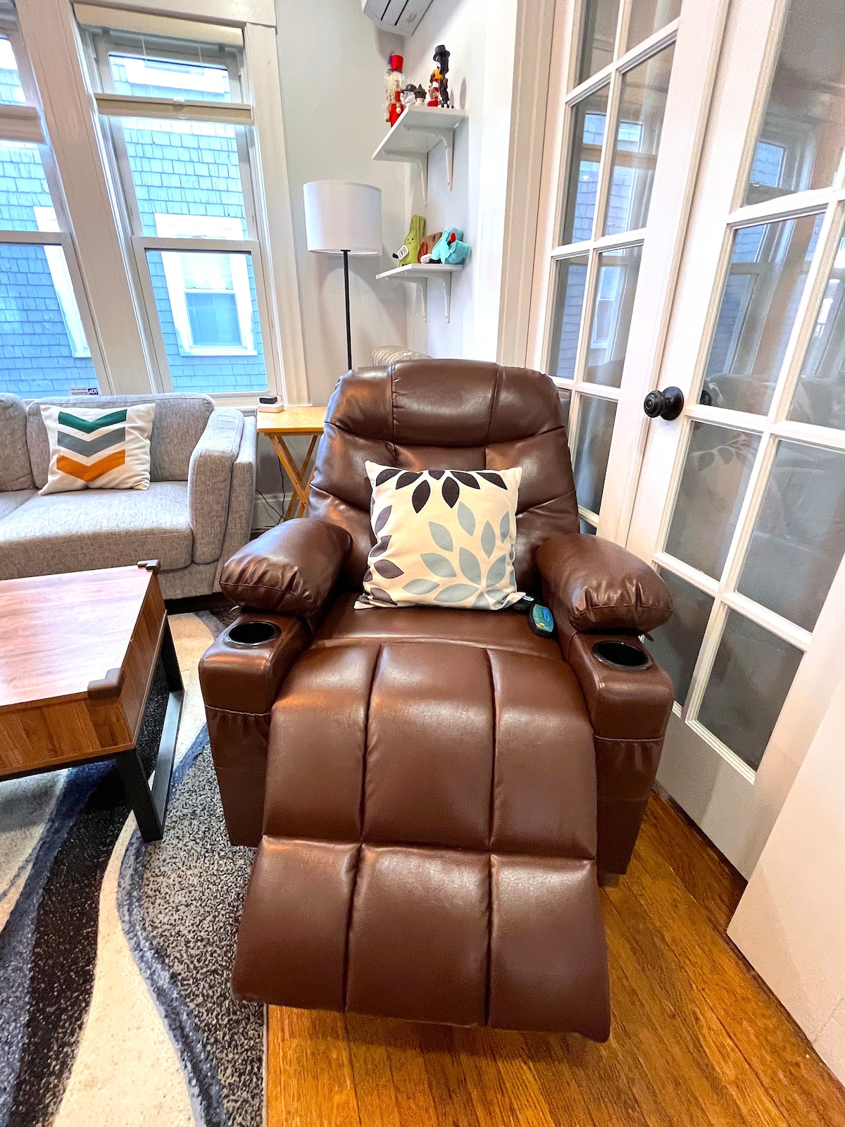 A recliner massage chair is positioned in a cozy living area, featuring brown leather upholstery. A decorative pillow with a floral pattern rests on the chair. Nearby, a light-colored sofa and a wooden coffee table can be seen, along with natural light streaming through tall windows.