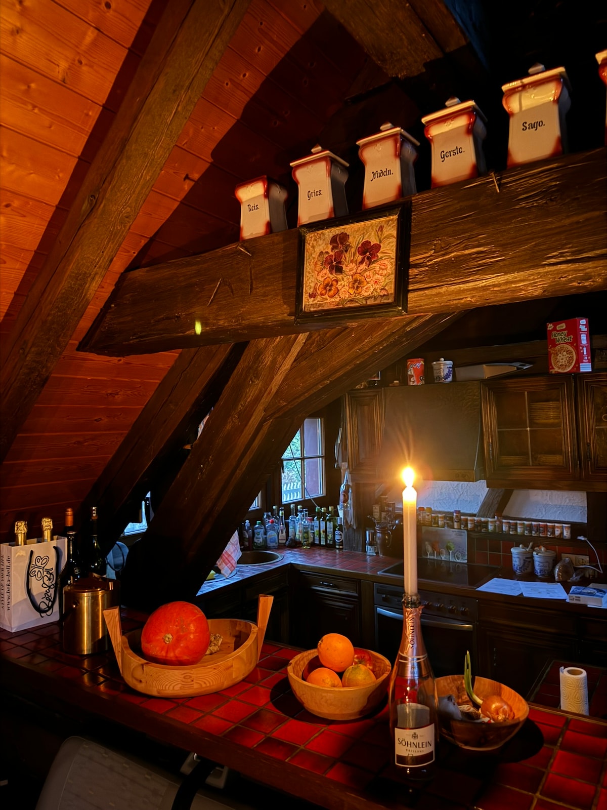 A rustic kitchen area features wooden beams and a rich brown color palette. Jars are neatly arranged on the shelf, and a candle casts a warm glow on the countertop, which holds a bowl of oranges, apples, and a bottle of sparkling drink.