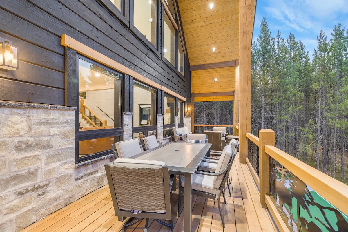 An outdoor deck is furnished with a large table surrounded by comfortable seating, creating an ideal space for dining or gatherings. The backdrop features tall, green pines, enhancing the sense of privacy and connection to nature.