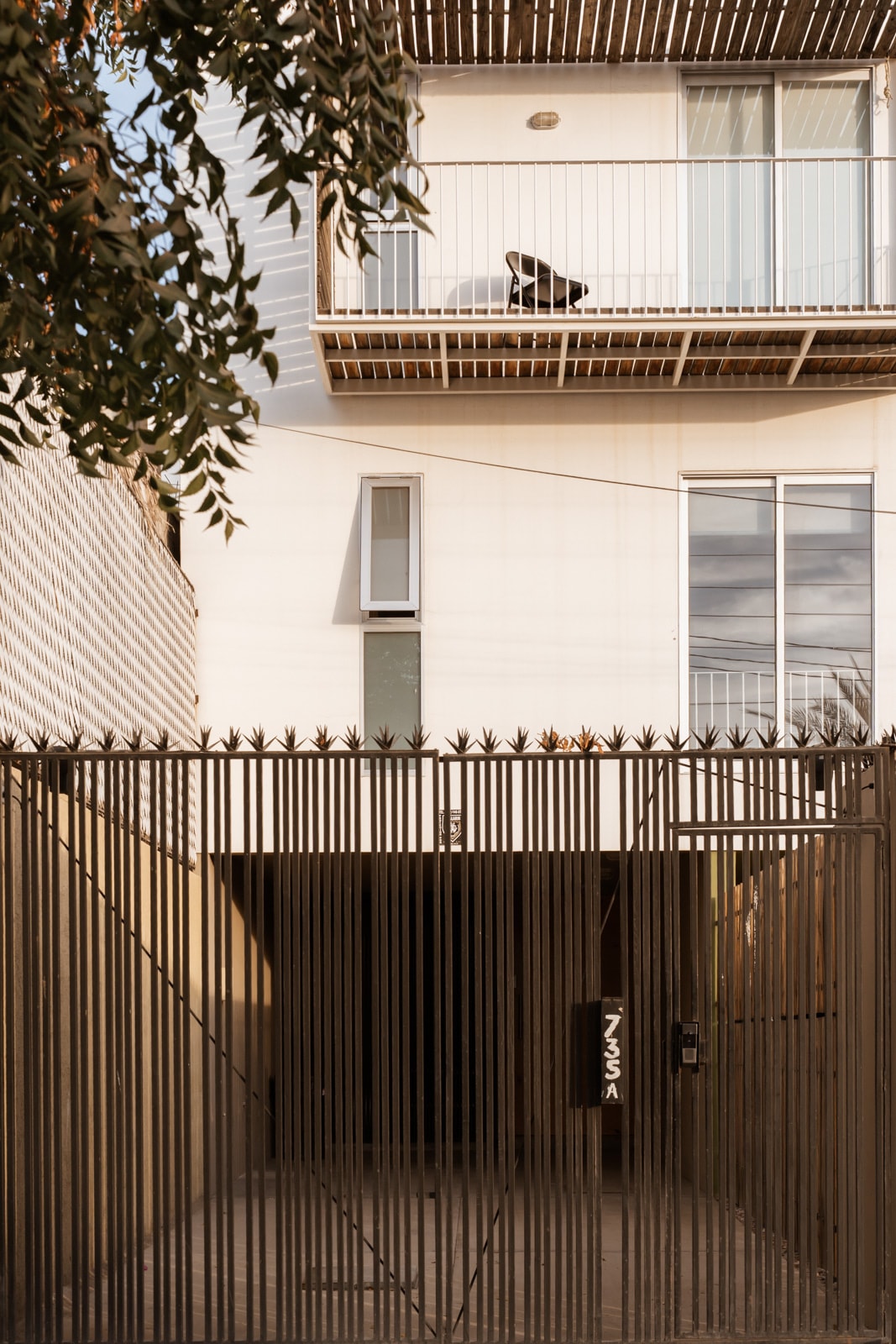 A modern townhouse entrance is depicted, featuring a gated entry with vertical bars. Above, a balcony is visible with a seating arrangement. The facade is minimalistic, showcasing large windows and neutral-colored walls, allowing natural light to illuminate the area.