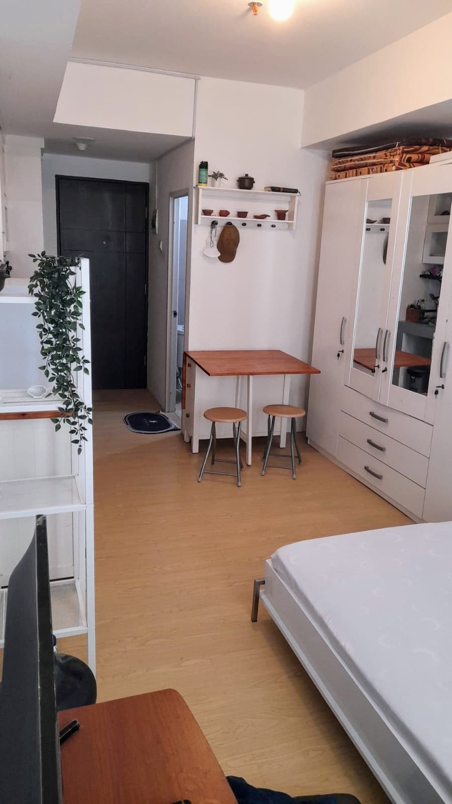 An interior view of a compact apartment is displayed, featuring a queen bed and an extra single bed. A small dining table with two chairs is positioned near a wall-mounted shelf, with white cabinetry providing storage. A TV is placed on a stand in the foreground.