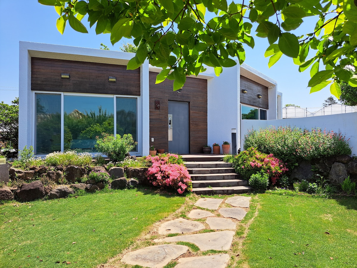 A modern, single-story structure is set amidst well-maintained gardens. Natural stone steps lead to a wooden front door, flanked by large windows that allow for ample natural light. Colorful flowers and greenery create a vibrant and inviting entrance.