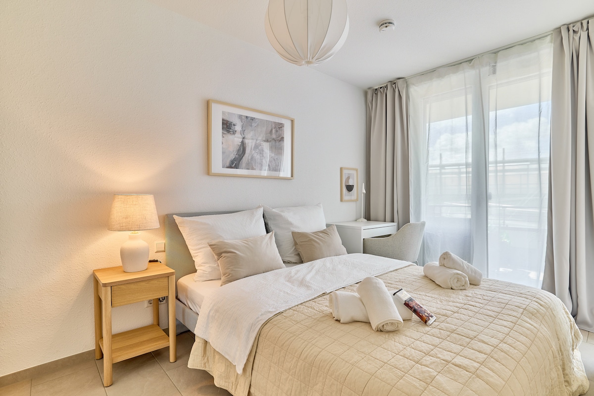 A serene bedroom is displayed, featuring a spacious bed adorned with neutral-colored bedding. A bedside table with a lamp is positioned next to the bed. Light flows in through large windows, complemented by soft curtains, creating a calming ambiance throughout the room.