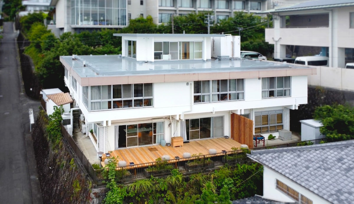 The three-story building is framed by greenery, highlighting its modern design. A wooden deck area is visible at the front, offering a space for outdoor relaxation. Large windows reflect natural light, creating an open and inviting appearance.