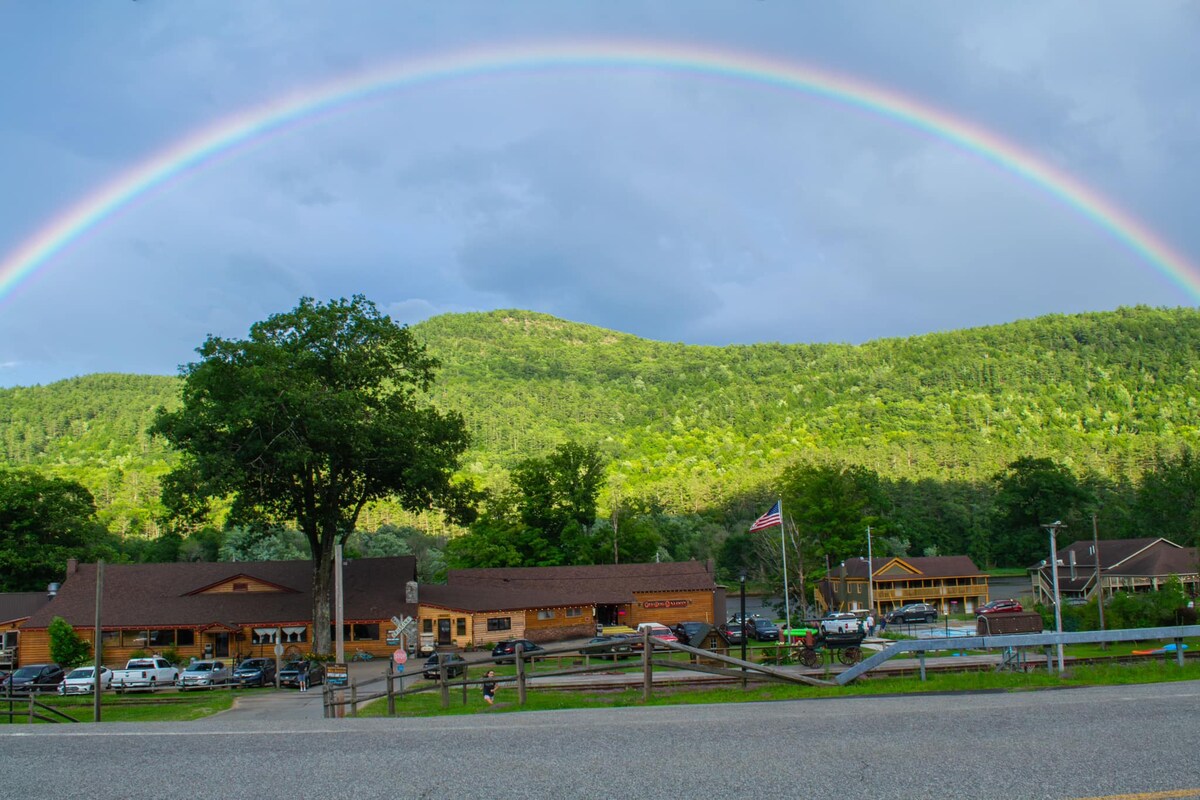 A scenic view captures the exterior of the lodge against a backdrop of lush green mountains, with a vibrant rainbow arching across the sky. Vehicles are parked in front, and an American flag is visibly displayed nearby.