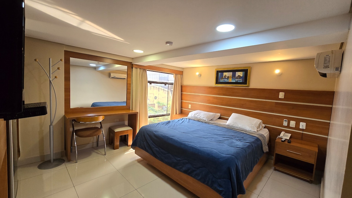 A well-lit sleeping area features a spacious bed with blue bedding. A wooden desk and chair are positioned near a large mirror, while natural light enters through a window dressed with light curtains. A flat-screen TV is mounted on the wall.