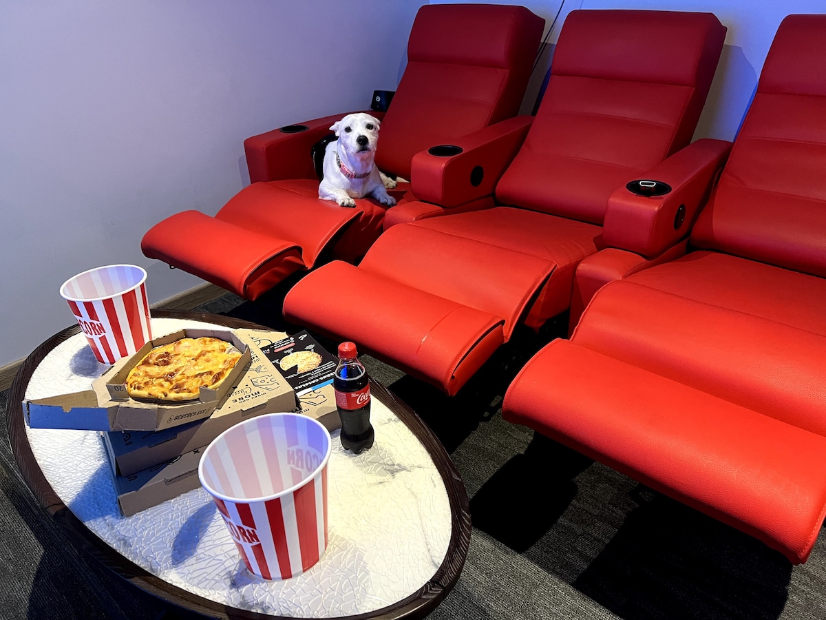 The image features a cozy movie watching area with three red reclining chairs positioned around a small table. A dog is seen sitting on one chair, while a snack table displays popcorn, pizza, and a bottle of soda, creating an inviting setting for relaxation.