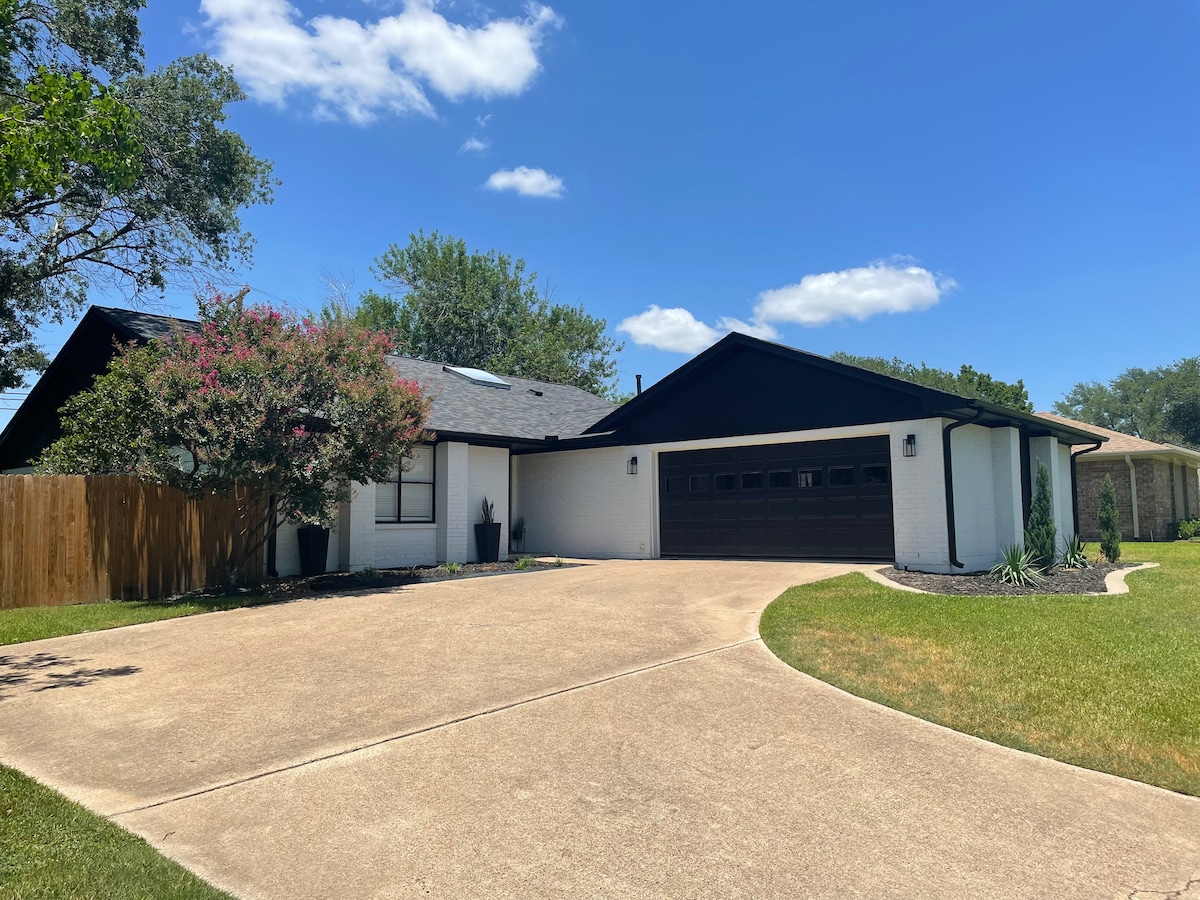 The exterior of a recently updated home is shown, featuring a well-maintained driveway with a double garage. Lush green grass borders the property, complemented by flowering shrubs along the side. A wooden fence provides privacy, with a clear blue sky and fluffy clouds in the background.