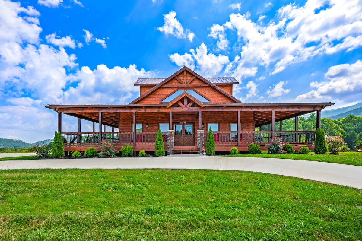 The custom-built lodge is presented with a wooden facade and a large covered porch. Lush greenery surrounds the property, complemented by flowering shrubs. The design incorporates tall gables and expansive windows, allowing natural light to enhance the inviting exterior on a sunny day.