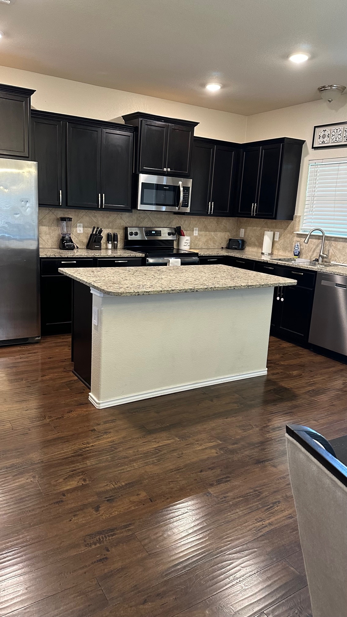 A modern kitchen features dark cabinetry and granite countertops. Stainless steel appliances, including a refrigerator and oven, create a sleek look. Ample natural light comes through the window, illuminating the open space and enhancing the inviting atmosphere of the room.