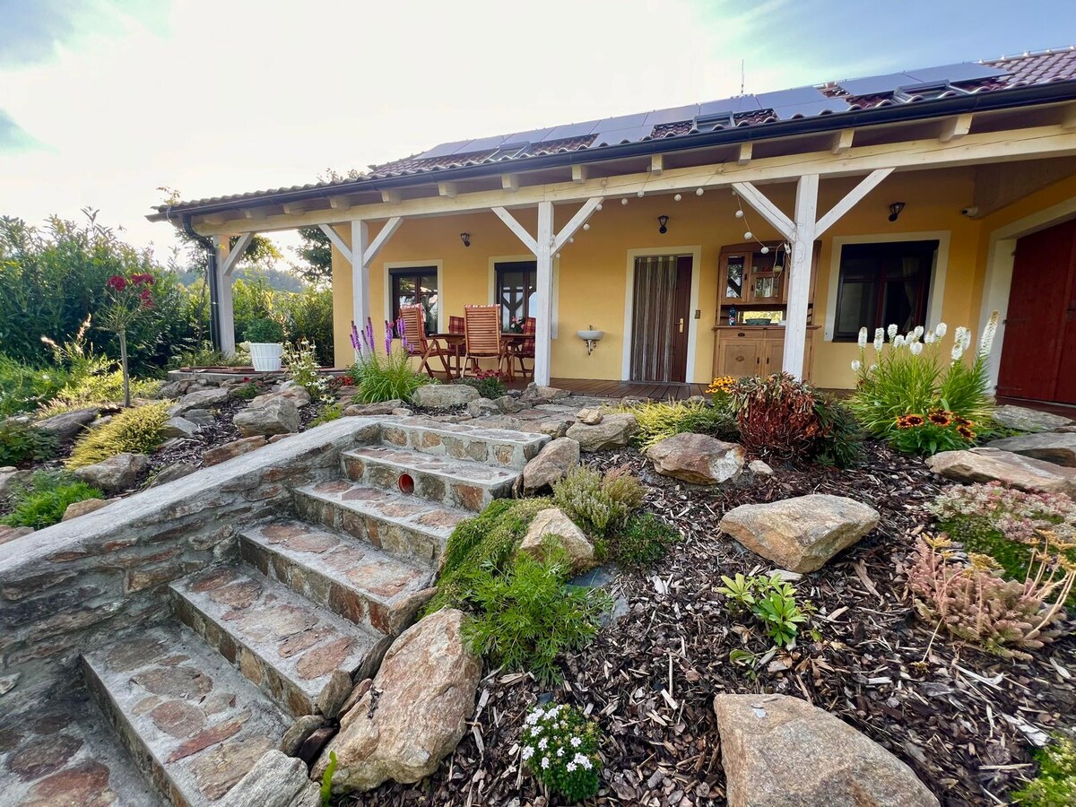 The exterior of the cozy chalet features a stone pathway leading to the entrance, surrounded by a variety of plants and flowers. Natural stone steps ascend from a landscaped garden, complemented by wooden seating positioned under the overhang.