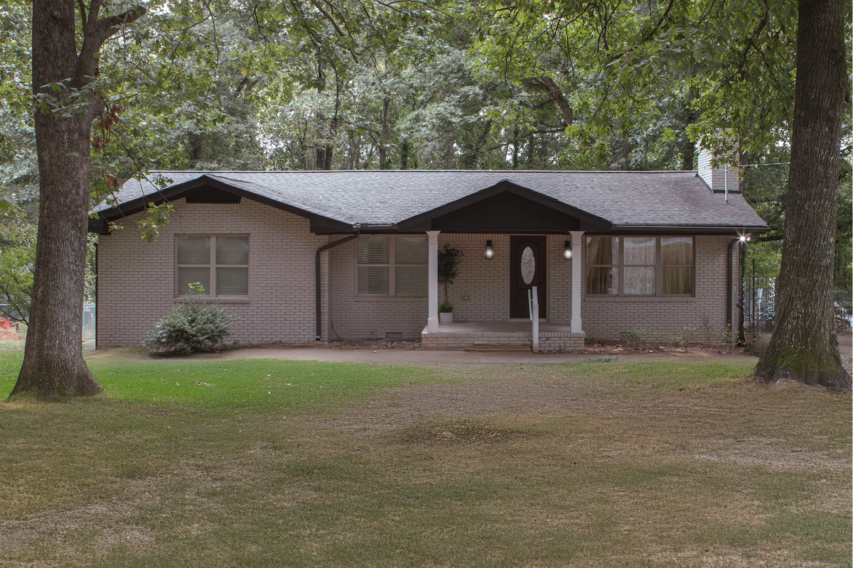 The exterior of a chic single-story residence is framed by tall trees, showcasing a light-colored brick facade. A welcoming front porch with a round door adds charm, while the manicured lawn is bordered by gravel pathways. The overall setting conveys a serene environment.