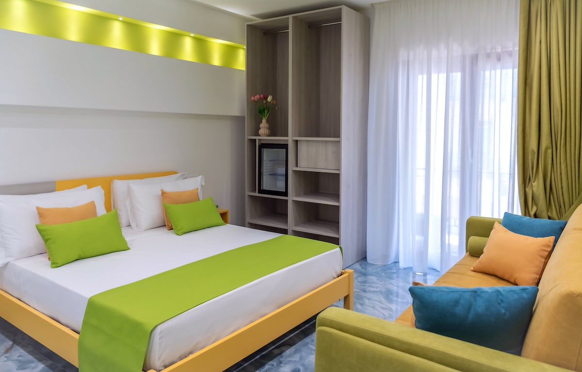 A spacious room is designed with bright accents in green and orange. A large bed, flanked by decorative pillows, is complemented by a cozy sofa. Natural light filters through sheer curtains, illuminating the room's modern furnishings and minimalist shelving.