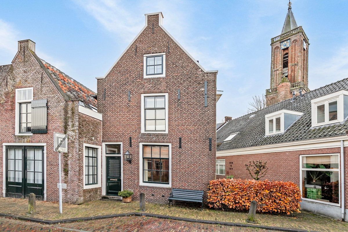 The exterior of a charming house features a mix of red brick and white window frames, complemented by a well-maintained garden with seasonal foliage. A clock tower is visible in the background, providing a historic context to the inviting streetscape.