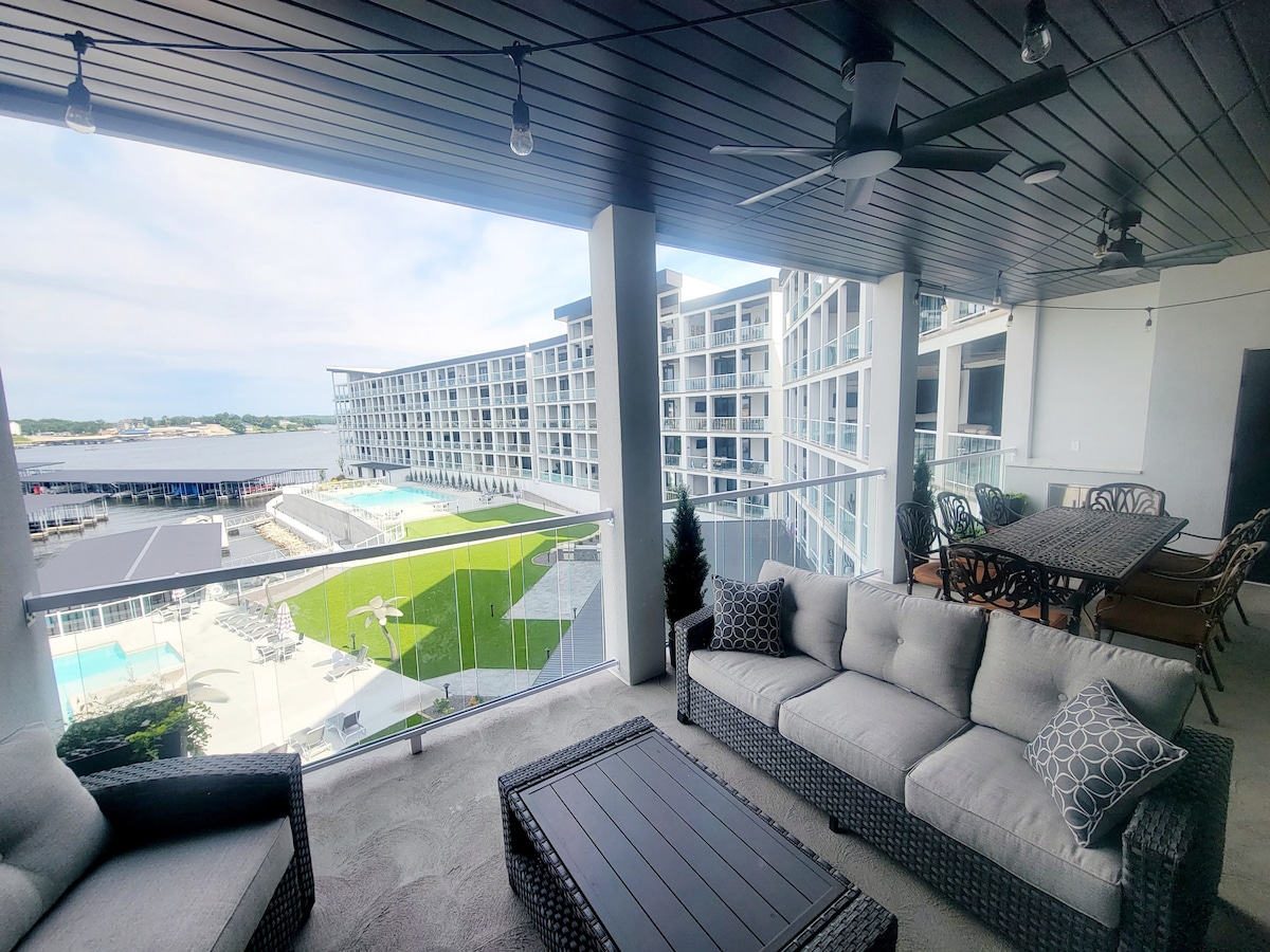 A spacious covered patio features comfortable seating arrangements with a sectional sofa and a coffee table. Dining tables provide additional seating. The area offers views of the surrounding lush green landscape and sparkling pools, creating a serene outdoor environment.
