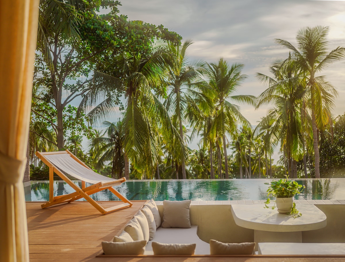 An outdoor seating area features a wooden lounge chair positioned beside an infinity pool. Lush palm trees surround the space, creating a natural backdrop. Soft cushions line a built-in seating area, inviting relaxation as sunlight filters through the trees.