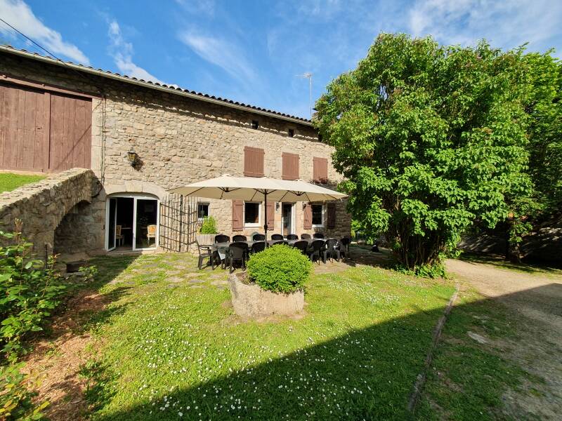 A spacious outdoor area features a rustic stone building with wooden shutters. A large patio is shaded by an umbrella, surrounded by a set of black chairs and a stone table. Lush greenery and blooming flowers enhance the inviting ambiance.